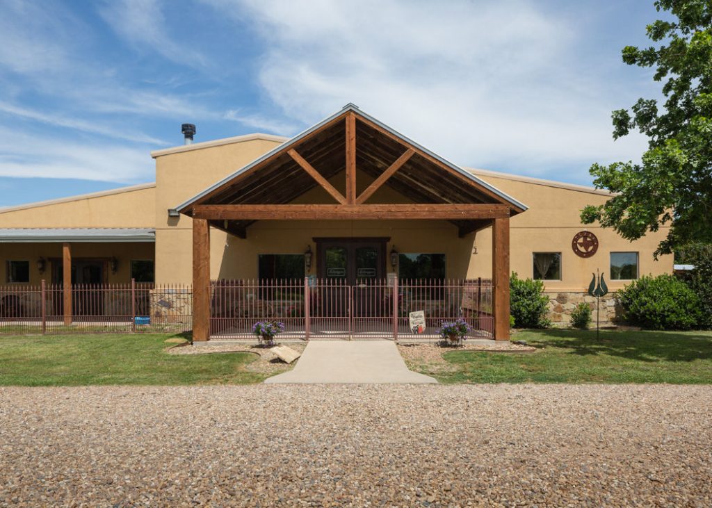 Building with a wooden pergola entrance, beige walls, gravel path, and landscaped lawn under a partly cloudy sky.