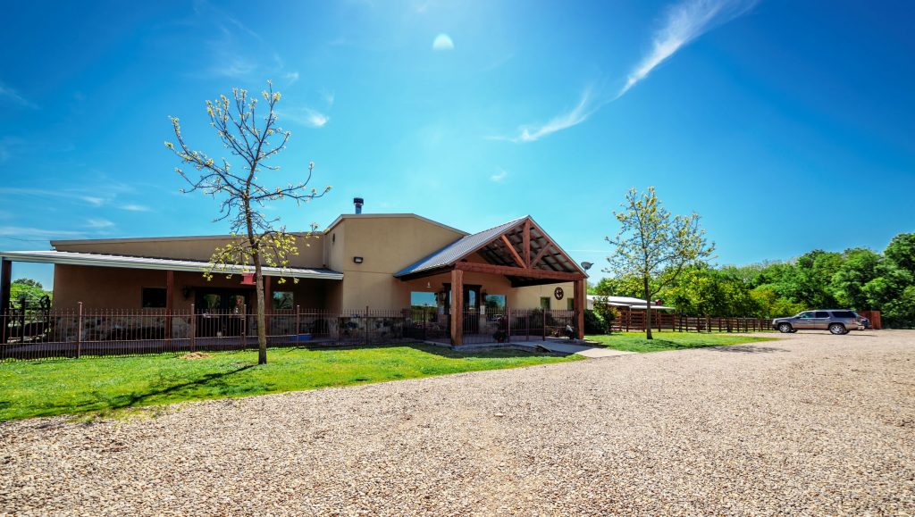 A single-story building with a gabled entrance, surrounded by trees and a gravel parking area under a clear blue sky.