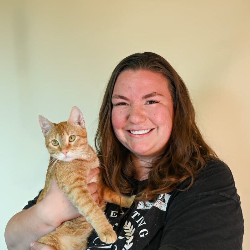 Person smiling while holding an orange tabby cat against a plain background.